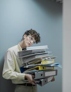 Young man with glasses carrying a stack of files looking tired. Office setting with gray wall.